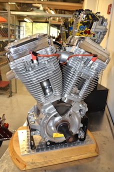 A close-up view of a V-twin motorcycle engine resting on a stand. The engine has shiny metallic surfaces with visible fins and red ignition wires. It appears to be a large, powerful engine designed for high performance.