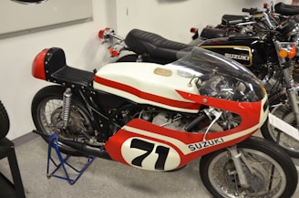 A vintage Suzuki racing motorcycle with a racing number 71 is displayed indoors. The bike features a red and white color scheme with a streamlined fairing and a transparent windscreen. Adjacent to it are other classic Suzuki motorcycles, showcasing similar retro designs.
