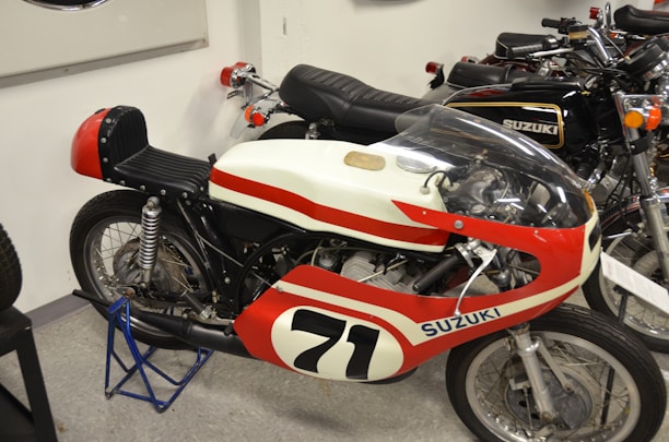 A vintage Suzuki racing motorcycle with a racing number 71 is displayed indoors. The bike features a red and white color scheme with a streamlined fairing and a transparent windscreen. Adjacent to it are other classic Suzuki motorcycles, showcasing similar retro designs.