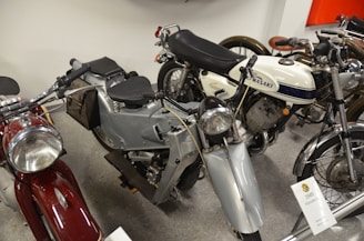 A collection of vintage motorcycles is displayed, including models with classic designs. Prominent details such as a white Kawasaki bike with blue accents and a silver Velocette with black seat stand out. The motorcycles are closely arranged in an indoor setting, suggesting a museum or exhibition environment.