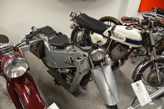 A collection of vintage motorcycles is displayed, including models with classic designs. Prominent details such as a white Kawasaki bike with blue accents and a silver Velocette with black seat stand out. The motorcycles are closely arranged in an indoor setting, suggesting a museum or exhibition environment.