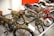 A collection of vintage motorcycles featuring distinctive designs and classic components. The motorcycles have a range of styles, including a green military-themed bike with saddlebags and a leather seat, and a red and white bike with a shiny exhaust pipe. The scene is set in an indoor museum-like environment with several other motorcycles visible in the background.