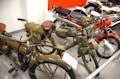 A collection of vintage motorcycles featuring distinctive designs and classic components. The motorcycles have a range of styles, including a green military-themed bike with saddlebags and a leather seat, and a red and white bike with a shiny exhaust pipe. The scene is set in an indoor museum-like environment with several other motorcycles visible in the background.