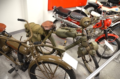 A collection of vintage motorcycles featuring distinctive designs and classic components. The motorcycles have a range of styles, including a green military-themed bike with saddlebags and a leather seat, and a red and white bike with a shiny exhaust pipe. The scene is set in an indoor museum-like environment with several other motorcycles visible in the background.
