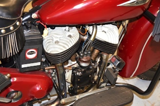 A close-up view of a vintage motorcycle engine featuring a red fuel tank with chrome accents and visible logos. The twin-cylinder engine shows metal components with black and silver finishes, surrounded by various wires and parts. The image captures the intricate design details and mechanical complexity.