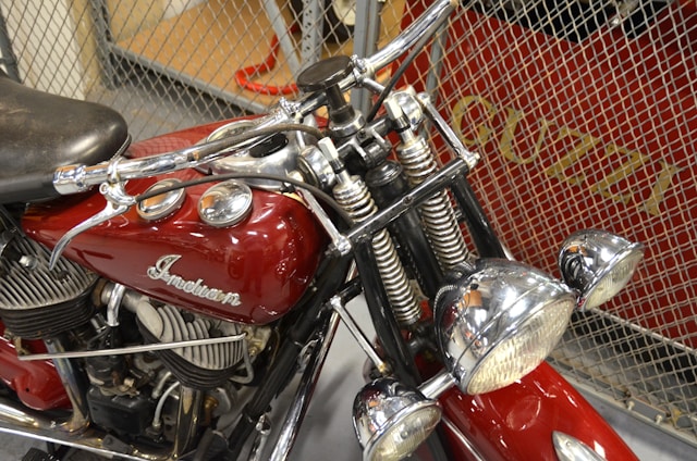 A vintage motorcycle features prominently with a red fuel tank bearing the word 'Indian'. The bike is equipped with chrome handlebars and twin chrome headlights. The intricate detail of the engine and the overall classic design suggest an era-specific craftsmanship. The background shows a metal wire mesh with some text partially visible.
