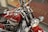 A vintage motorcycle features prominently with a red fuel tank bearing the word 'Indian'. The bike is equipped with chrome handlebars and twin chrome headlights. The intricate detail of the engine and the overall classic design suggest an era-specific craftsmanship. The background shows a metal wire mesh with some text partially visible.
