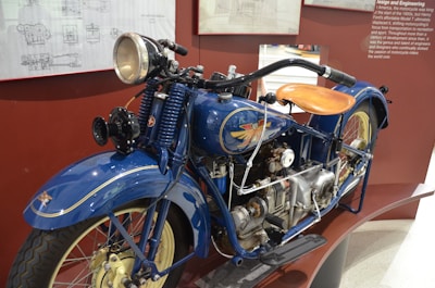 A vintage blue motorcycle with intricate mechanical details is displayed against a backdrop of informational panels. The motorcycle features a prominent headlight, a curved leather seat, and an exposed engine. The design includes classic elements such as wire-spoked wheels and detailed insignia on the fuel tank.