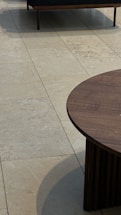 A modern chair with a dark cushion is partially visible on a light tiled floor. A circular wooden coffee table with vertical slats is prominently displayed in the foreground.