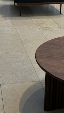 A modern chair with a dark cushion is partially visible on a light tiled floor. A circular wooden coffee table with vertical slats is prominently displayed in the foreground.