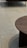 A modern chair with a dark cushion is partially visible on a light tiled floor. A circular wooden coffee table with vertical slats is prominently displayed in the foreground.