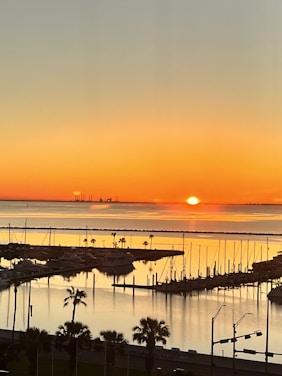 Sunset over sailboats glowing orange near a calm waterfront patio with fountains and chairs.