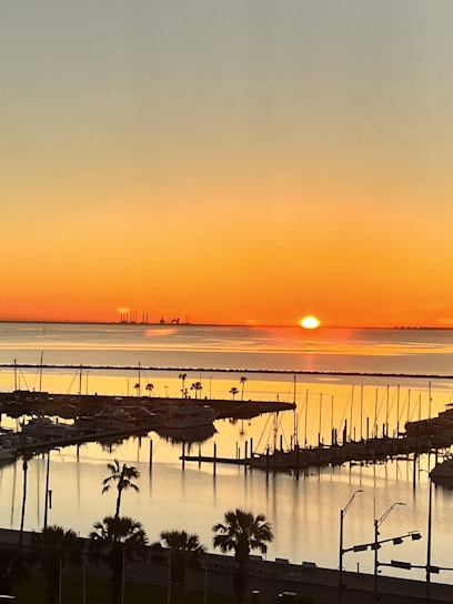 Sunset over sailboats glowing orange near a calm waterfront patio with fountains and chairs.