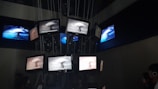 A dark room containing multiple screens suspended and arranged in a clustered formation. The screens display various images of a nuclear explosion, with some screens showing the explosion in black and white and others in color. Lines and cables are visible, supporting the screens from above, while people are seen observing the display.