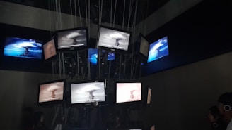 A dark room containing multiple screens suspended and arranged in a clustered formation. The screens display various images of a nuclear explosion, with some screens showing the explosion in black and white and others in color. Lines and cables are visible, supporting the screens from above, while people are seen observing the display.