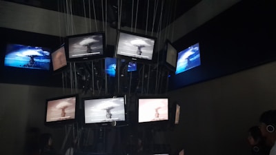 A dark room containing multiple screens suspended and arranged in a clustered formation. The screens display various images of a nuclear explosion, with some screens showing the explosion in black and white and others in color. Lines and cables are visible, supporting the screens from above, while people are seen observing the display.