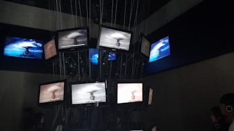A dark room containing multiple screens suspended and arranged in a clustered formation. The screens display various images of a nuclear explosion, with some screens showing the explosion in black and white and others in color. Lines and cables are visible, supporting the screens from above, while people are seen observing the display.