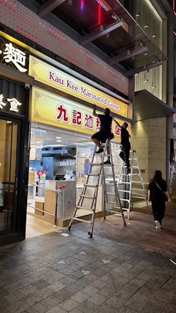 Two people are standing on ladders, adjusting or installing the signage for a restaurant called 'Kau Kee Marinated Goose'. The scene is set at night, with one person reaching up to the sign and another holding a tool. The restaurant interior is partially visible, showcasing a well-lit, modern design with kitchen equipment.