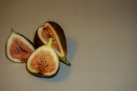 A close-up of three ripe figs, with two halves exposing their pinkish-red flesh and a whole fig in the background on a neutral surface.