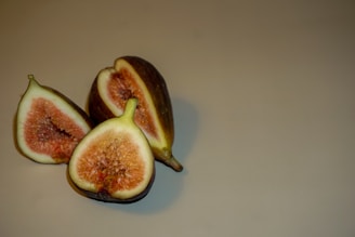 A close-up of three ripe figs, with two halves exposing their pinkish-red flesh and a whole fig in the background on a neutral surface.