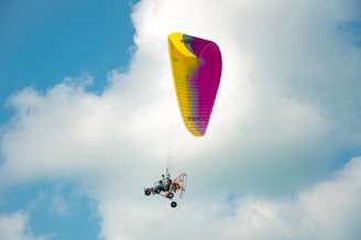 A powered paraglider is soaring through the sky against a backdrop of fluffy clouds. The wing is bright yellow with some pink areas, and the pilot is seated below with the motor visible.