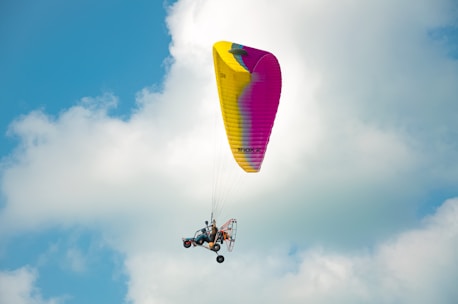 A powered paraglider is soaring through the sky against a backdrop of fluffy clouds. The wing is bright yellow with some pink areas, and the pilot is seated below with the motor visible.