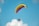 A paraglider with a brightly colored canopy, featuring hues of yellow and pink, against a clear blue sky. A person is seated in a powered parachute below the canopy, with equipment and a propeller visible.