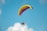 A paraglider with a brightly colored canopy, featuring hues of yellow and pink, against a clear blue sky. A person is seated in a powered parachute below the canopy, with equipment and a propeller visible.
