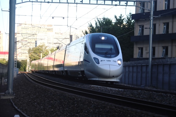 A sleek high-speed train travels on a curved railway track through an urban area, with buildings and trees in the background.