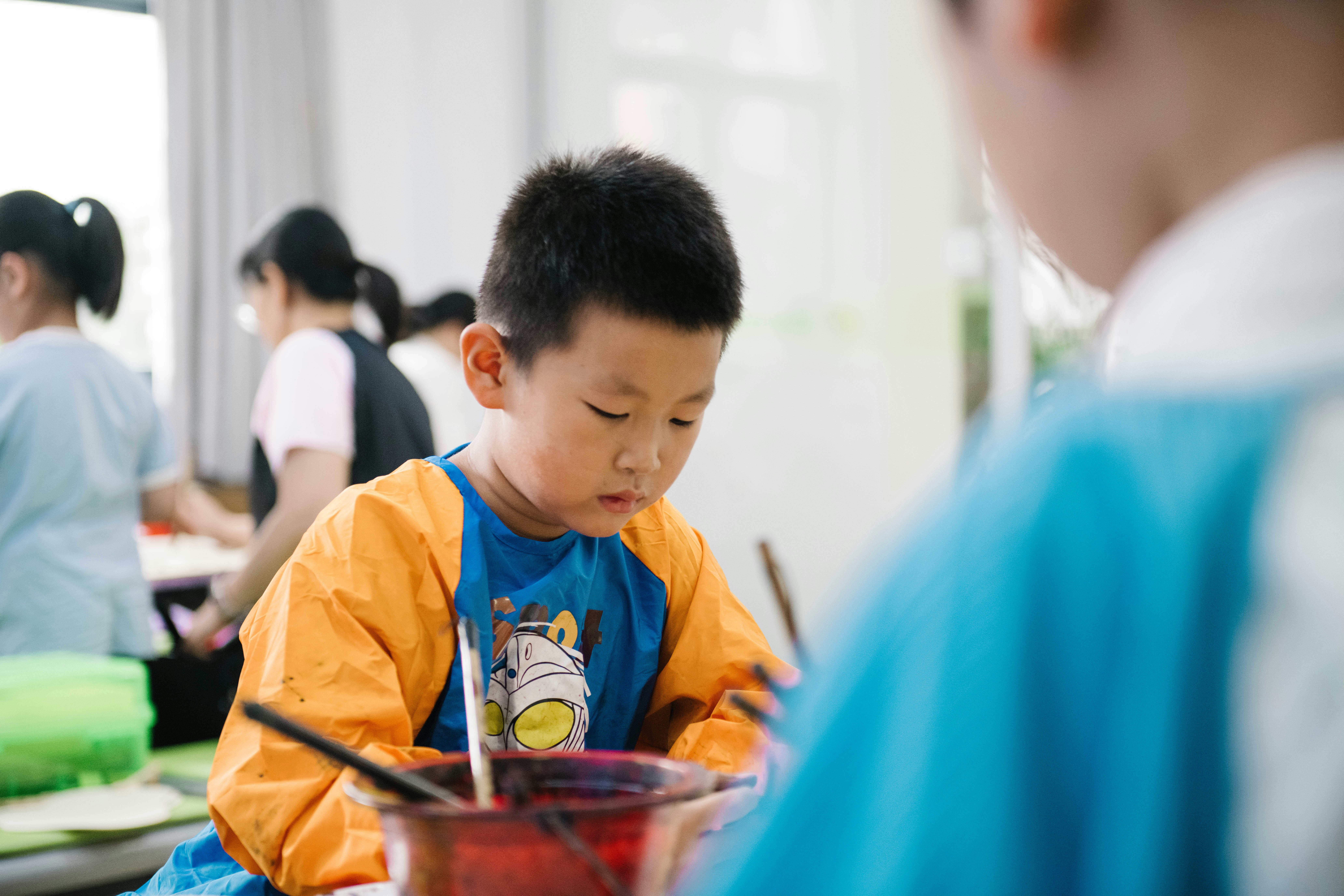 A young child is focused on an activity, wearing a colorful shirt with long sleeves, possibly for painting. Other children and some adults are in the background, likely engaged in similar activities in a brightly lit room.