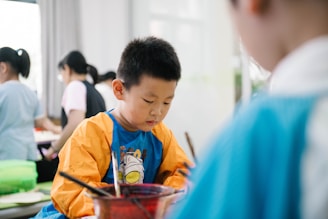 A young child is focused on an activity, wearing a colorful shirt with long sleeves, possibly for painting. Other children and some adults are in the background, likely engaged in similar activities in a brightly lit room.