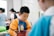 A young child is focused on an activity, wearing a colorful shirt with long sleeves, possibly for painting. Other children and some adults are in the background, likely engaged in similar activities in a brightly lit room.