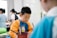 A young child is focused on an activity, wearing a colorful shirt with long sleeves, possibly for painting. Other children and some adults are in the background, likely engaged in similar activities in a brightly lit room.