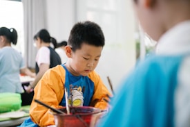 A young child is focused on an activity, wearing a colorful shirt with long sleeves, possibly for painting. Other children and some adults are in the background, likely engaged in similar activities in a brightly lit room.