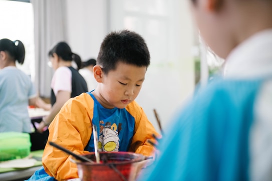 A young child is focused on an activity, wearing a colorful shirt with long sleeves, possibly for painting. Other children and some adults are in the background, likely engaged in similar activities in a brightly lit room.