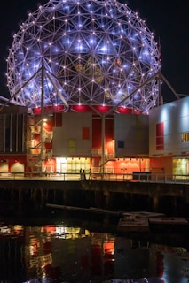 A geodesic dome illuminated at night, with a lattice structure of metal and lights creating a stunning architectural display. The building's facade is accented with red and white panels, and the entire structure is reflected in the calm water below, enhancing the visual symmetry and shine.