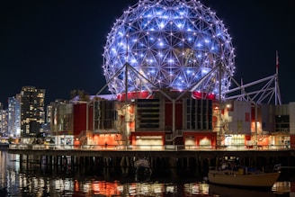 A geodesic dome structure beautifully illuminated with blue and white lights stands prominently against the night sky. Surrounding the dome is a modern, architectural building with red accents, situated by a waterfront. In the background, city skyscrapers are visible, their lights reflecting off the water.