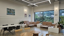 A modern, spacious lounge area with several brown leather sofas arranged around low coffee tables. The room has a white and beige color scheme with large windows providing a view of greenery outside. There is a white dining table with dark chairs to the left and some art pieces hanging on the walls. Several potted plants add a touch of nature to the decor.