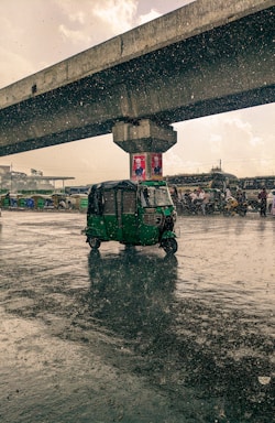 A green auto rickshaw drives through a wet road as rain pours heavily. Overhead, a concrete bridge spans the scene, with vehicles and people visible in the background. The surface of the road reflects the rain, creating a dramatic effect.