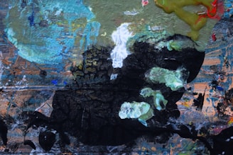 A vibrant abstract painting featuring a mix of textures and colors, including swirling areas of green, blue, and black. The composition includes a variety of brush strokes and layered paint applications, resulting in an intricate interplay of light and shadow. Some areas appear more chaotic with splashes of color, while others have a more structured, layered texture.