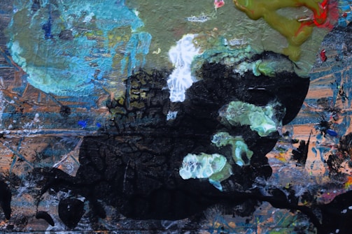 A vibrant abstract painting featuring a mix of textures and colors, including swirling areas of green, blue, and black. The composition includes a variety of brush strokes and layered paint applications, resulting in an intricate interplay of light and shadow. Some areas appear more chaotic with splashes of color, while others have a more structured, layered texture.