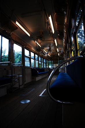 The image depicts the interior of an empty vintage tram or bus with wooden flooring and metal handrails. It has large windows on either side, allowing natural light to illuminate the space. Overhead lights are turned on, casting a warm glow. The seating is upholstered in blue fabric and the surroundings appear calm and quiet.