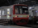 A red and white commuter train is stationed at an indoor platform. The train has open doors with a person in a yellow uniform visible inside. The platform has benches and a sign that reads 'Tanjung Priok'. The station's architecture features large arched windows and a steel roof structure.
