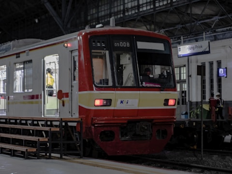 A red and white commuter train is stationed at an indoor platform. The train has open doors with a person in a yellow uniform visible inside. The platform has benches and a sign that reads 'Tanjung Priok'. The station's architecture features large arched windows and a steel roof structure.