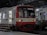 A red and white commuter train is stationed at an indoor platform. The train has open doors with a person in a yellow uniform visible inside. The platform has benches and a sign that reads 'Tanjung Priok'. The station's architecture features large arched windows and a steel roof structure.