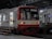 A red and white commuter train is stationed at an indoor platform. The train has open doors with a person in a yellow uniform visible inside. The platform has benches and a sign that reads 'Tanjung Priok'. The station's architecture features large arched windows and a steel roof structure.