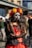 A person dressed in an elaborate costume with a red and black dress and intricate lace detailing. The face is painted with Day of the Dead motifs, accented with a crown of bright red and yellow flowers. Long black gloves with a spiderweb pattern cover the arms, and a black veil covers the head.
