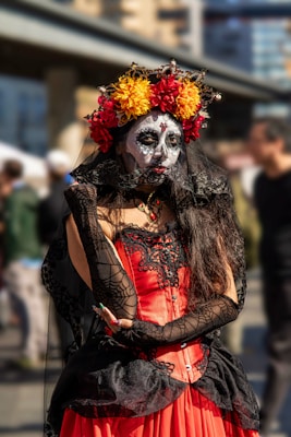 A person dressed in an elaborate costume with a red and black dress and intricate lace detailing. The face is painted with Day of the Dead motifs, accented with a crown of bright red and yellow flowers. Long black gloves with a spiderweb pattern cover the arms, and a black veil covers the head.