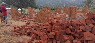 A construction site featuring unfinished brick walls and a large pile of red bricks in the foreground. A person wearing a patterned headscarf and colorful clothing stands to the left side, observing the site. The background shows grassy areas, sparse trees, and hills under a cloudy sky.