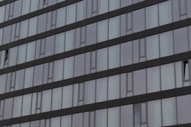 A modern building façade with a repeating pattern of rectangular windows and sleek, dark-colored frames. Some windows are slightly ajar, adding variation to the otherwise uniform structure. The reflections on the glass give a sense of depth and contemporary design.